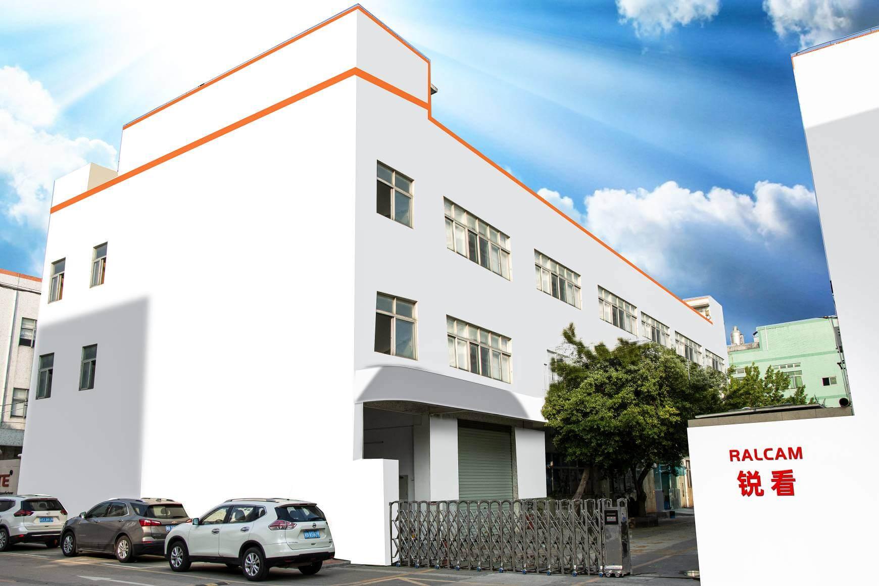 White industrial building with RALCAM signage and parked cars under blue sky with clouds